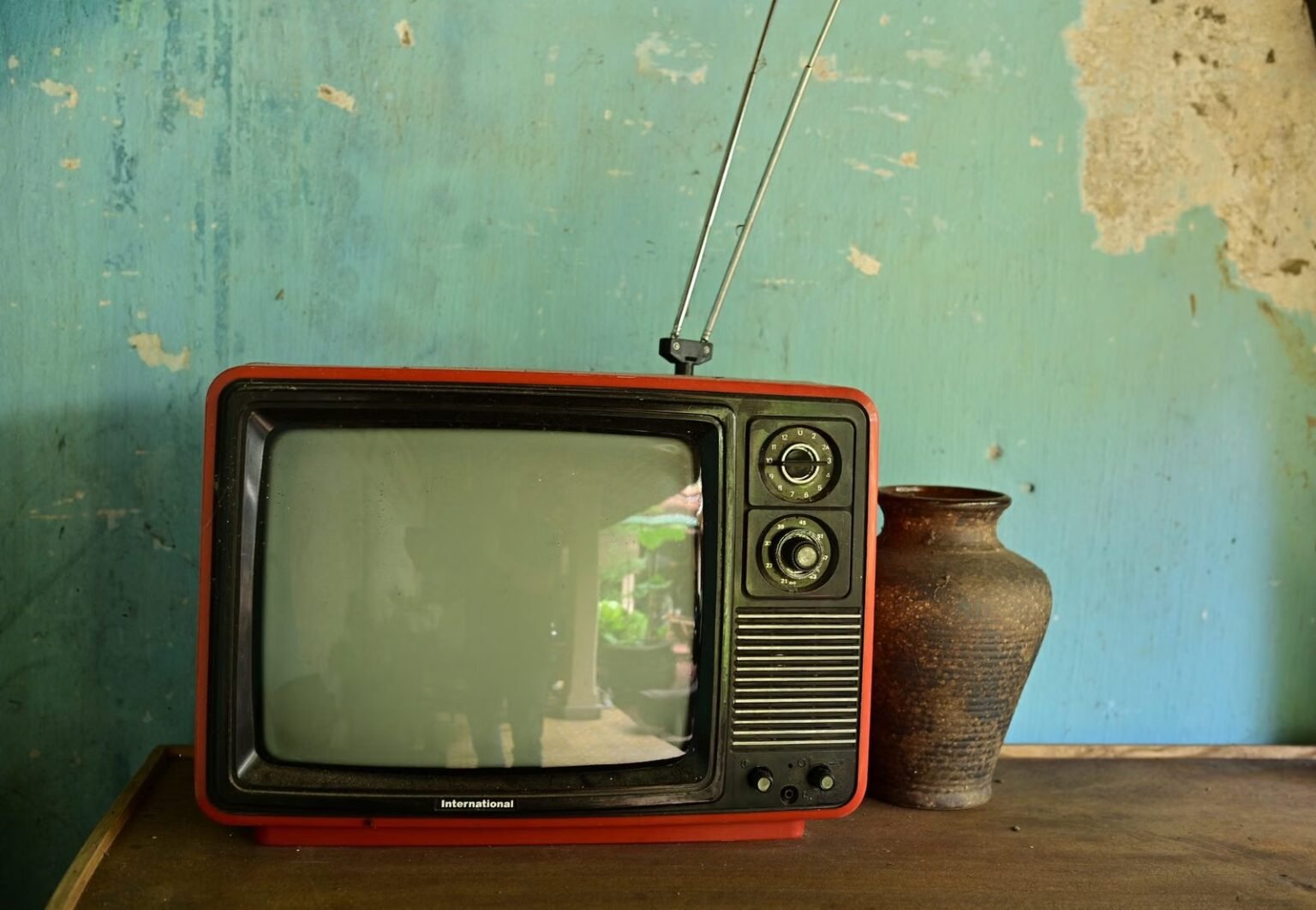 A vintage television with antenna beside a ceramic vase on a worn wooden surface against a rustic wall.