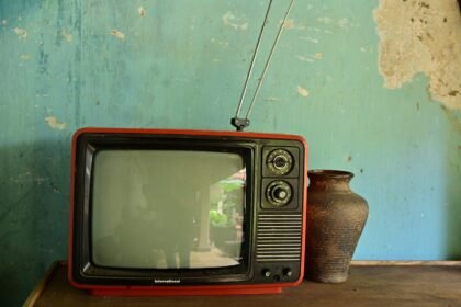 A vintage television with antenna beside a ceramic vase on a worn wooden surface against a rustic wall.