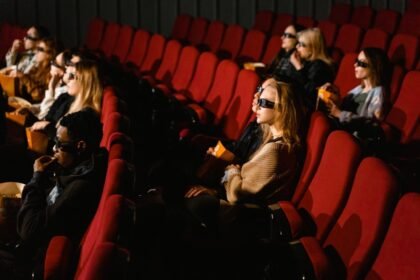 People wearing 3D glasses and eating popcorn in a cinema with red chairs.