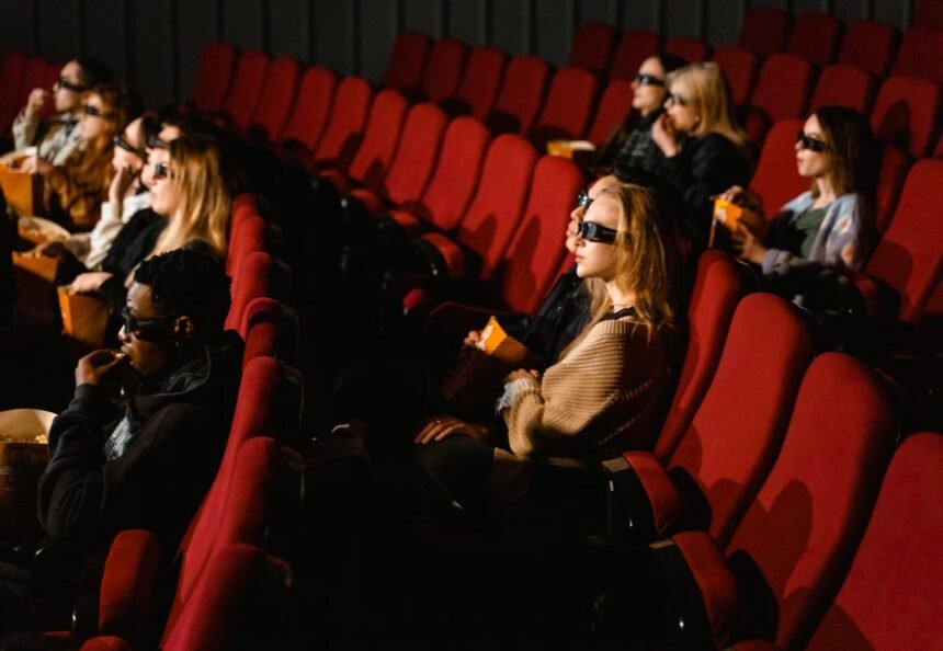 People wearing 3D glasses and eating popcorn in a cinema with red chairs.