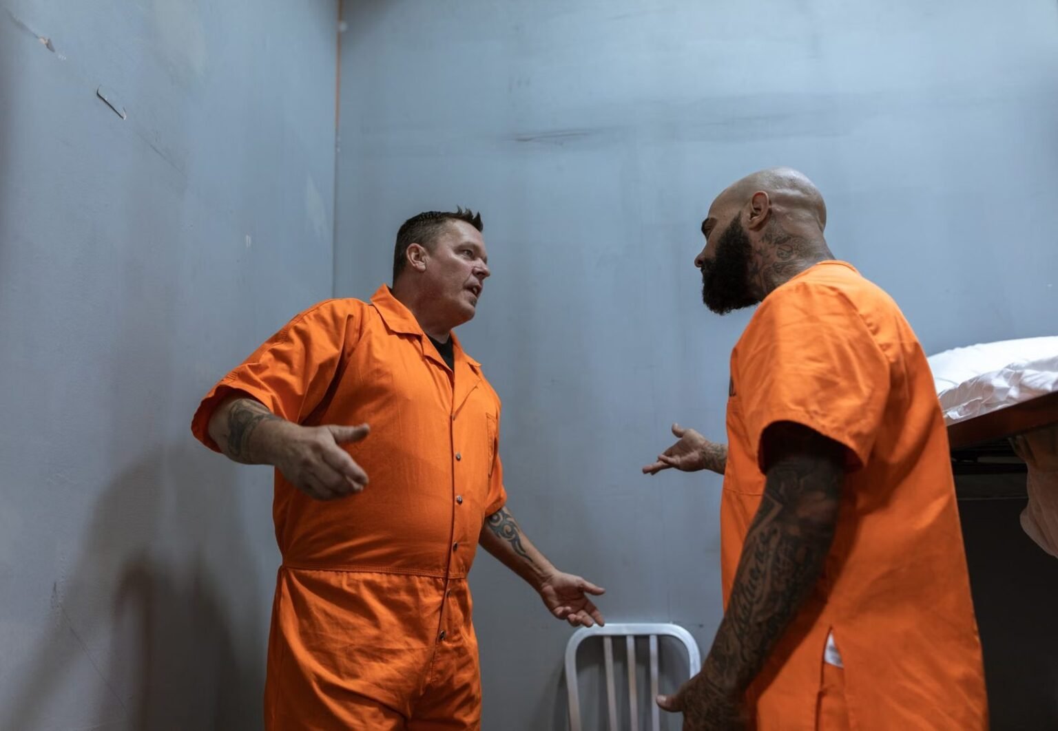 Two male prisoners in orange jumpsuits talking inside a prison cell.