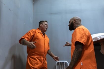 Two male prisoners in orange jumpsuits talking inside a prison cell.