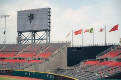 A modern stadium featuring red and gray seating and international flags, under a cloudy sky.