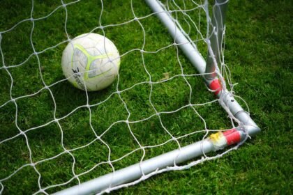 Close-up image of a soccer ball resting in a goal net on a vibrant green grass field.